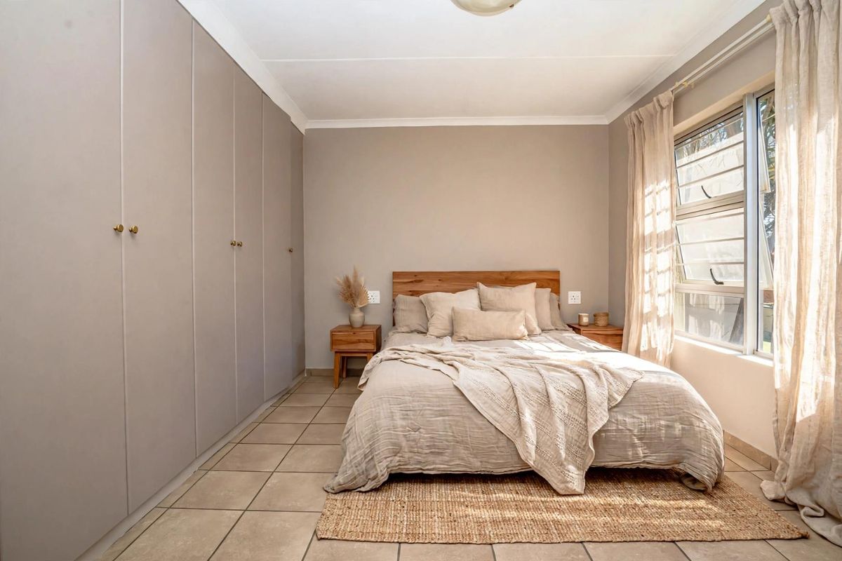 A well-maintained bedroom featuring neutral tones, extensive built-in cupboard storage, tiled flooring, and natural light from a large window.