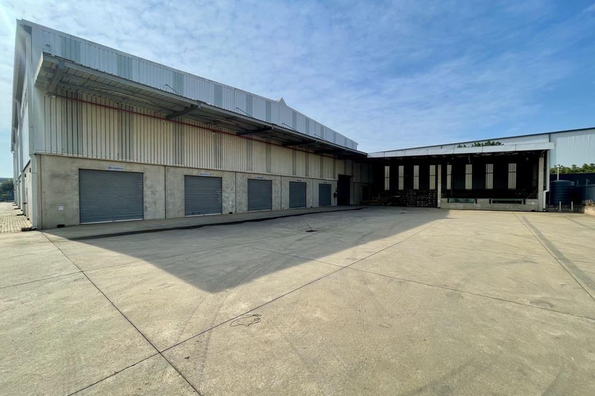 Industrial warehouse exterior featuring a large concrete yard, multiple roller shutter doors, and a covered loading bay area.