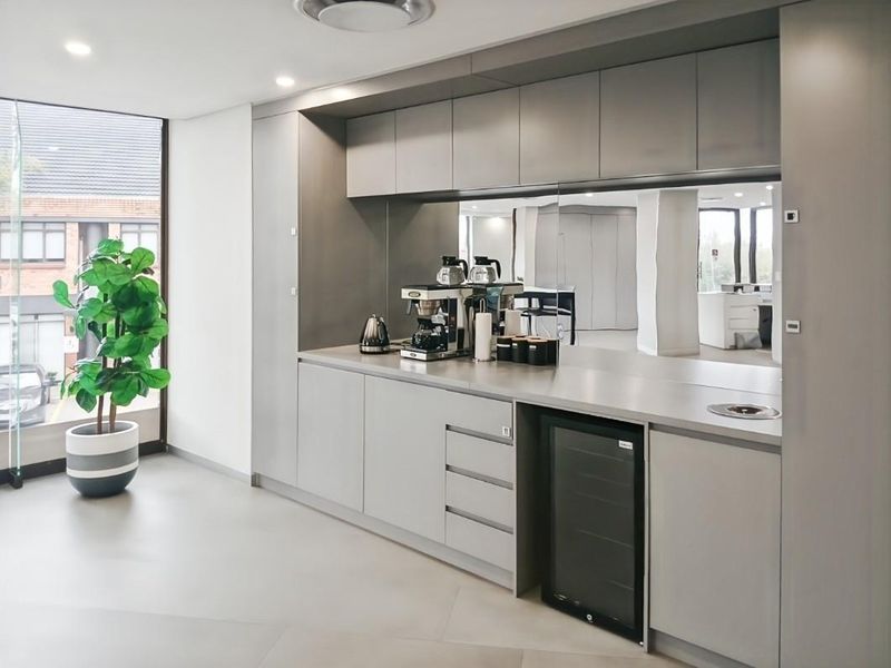 Modern office kitchenette featuring sleek grey cabinetry, a mirrored splashback, integrated beverage fridge, and recessed lighting, creating a clean and professional break area.