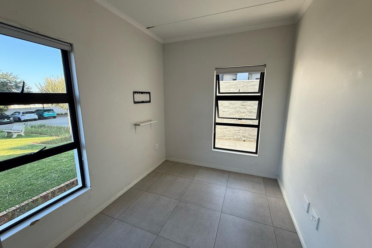 A bright, empty bedroom featuring neutral tiled flooring, large windows with black frames, and wall-mounted electrical and television points.