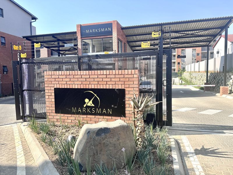The secure entrance to The Marksman apartment complex, featuring a brick facade, electric fencing, and a gated driveway.