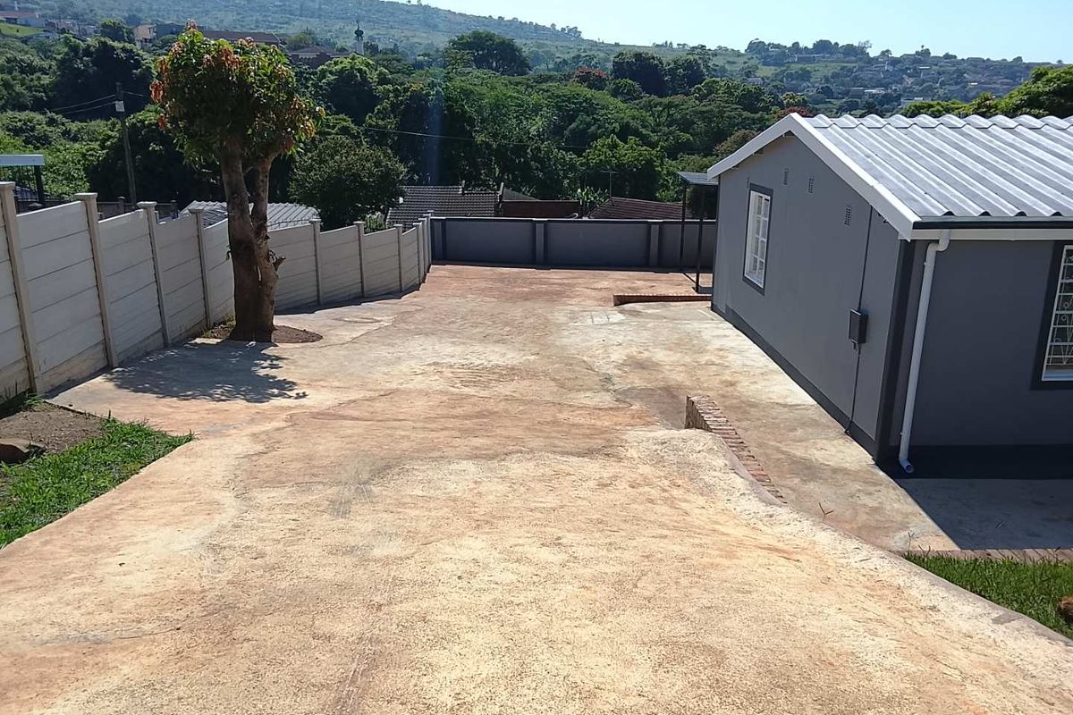 A spacious rear exterior area featuring a large concrete driveway, enclosed by precast boundary walls, with views of the surrounding landscape.