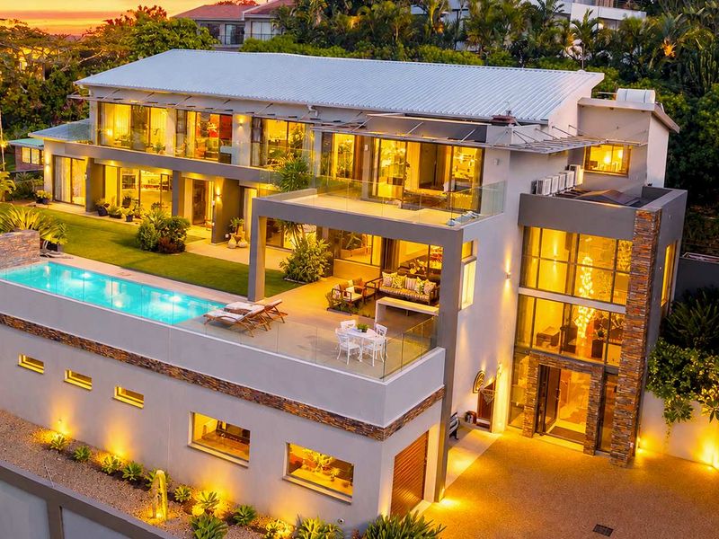 Stunning aerial view of a modern multi-level luxury residence featuring a swimming pool, expansive terraces, stone accents, and beautifully landscaped grounds at dusk.