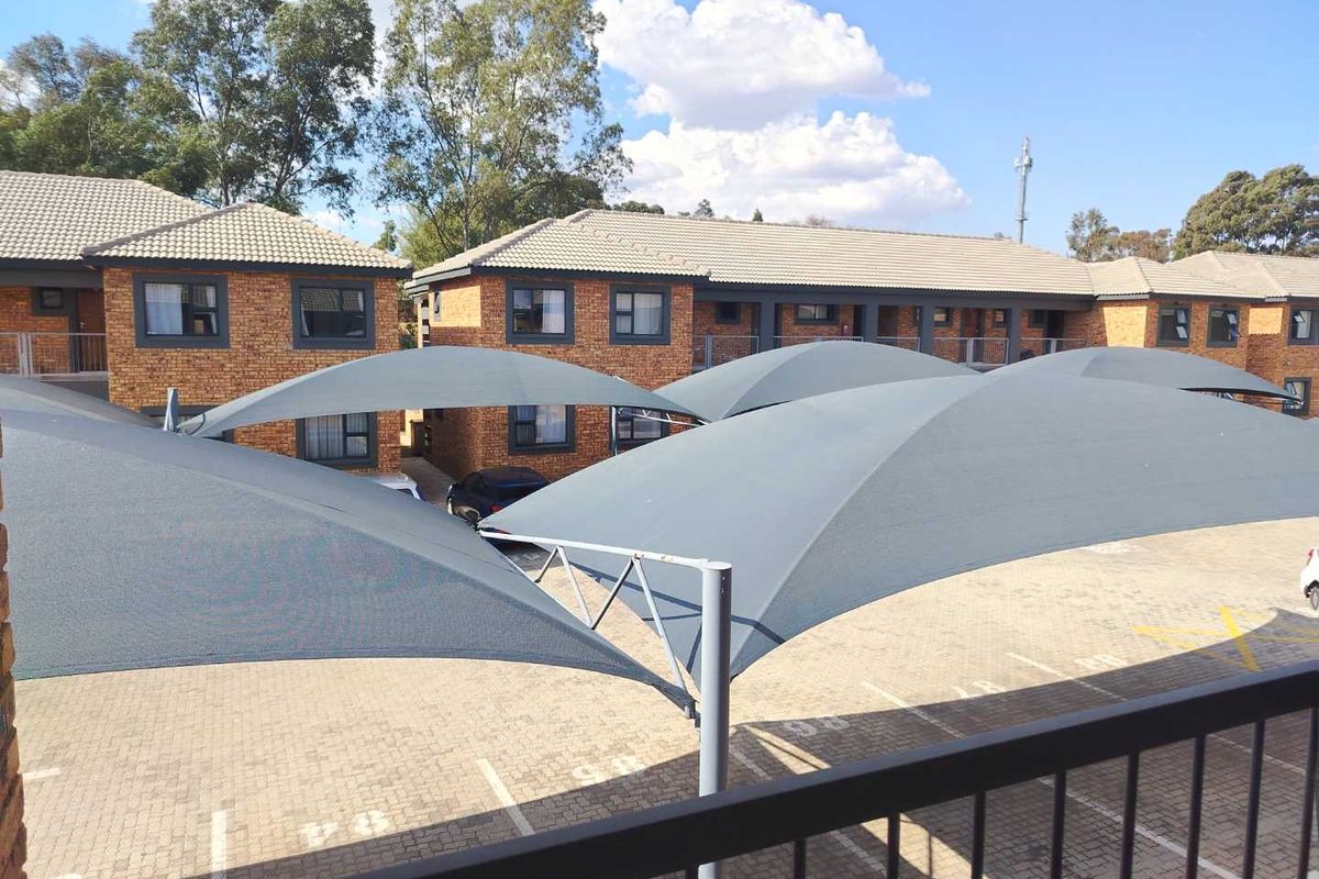 View of the apartment complex exterior featuring face brick buildings, a paved parking area with shade sail structures, and a metal balcony railing.