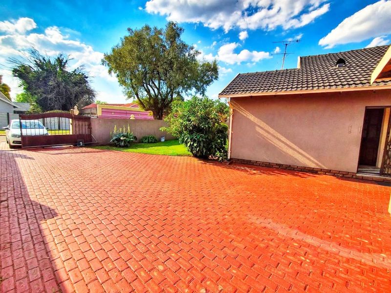 Spacious front exterior featuring a large paved driveway, secure metal gate, and a well-maintained garden area with established trees.