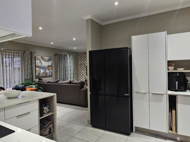 Modern open-plan kitchen featuring sleek white cabinetry, stone countertops, and integrated appliances, flowing seamlessly into a comfortable living area with tiled floors.