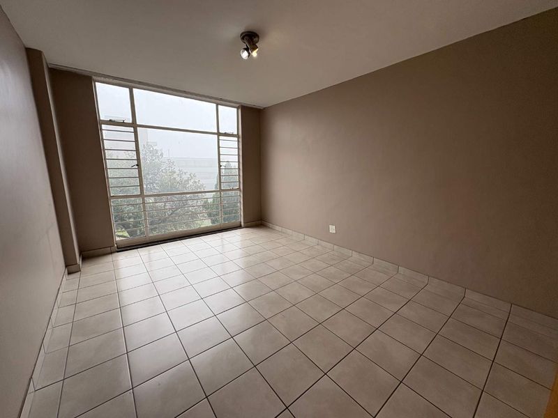An empty, neutrally painted living area featuring large metal-framed windows that provide ample natural light and tiled flooring throughout.