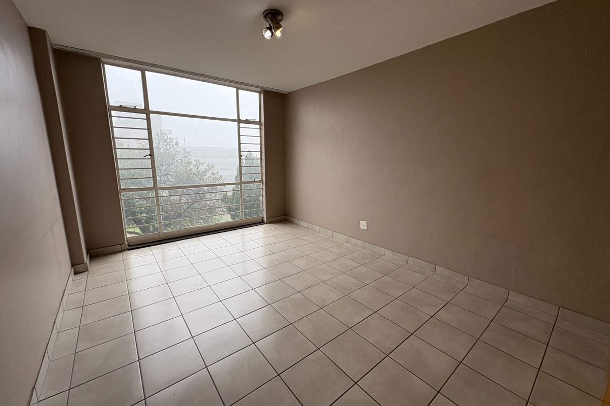 An empty, neutrally painted living area featuring large metal-framed windows that provide ample natural light and tiled flooring throughout.