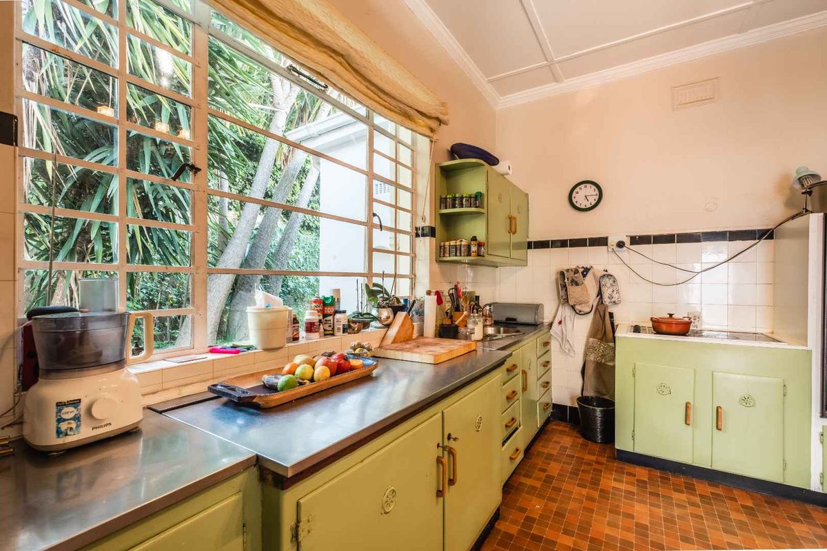 A vintage-style kitchen featuring original green cabinetry, stainless steel countertops, and large windows overlooking the garden, offering significant potential for a modern renovation.