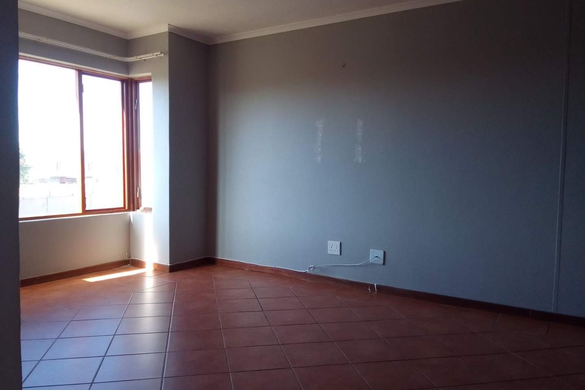 A bright, empty living room featuring terracotta floor tiles, neutral grey walls, and large windows providing natural light.