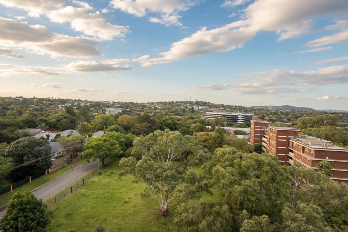 An elevated aerial view showcasing the surrounding neighbourhood, lush greenery, and nearby residential apartment buildings under a clear blue sky.