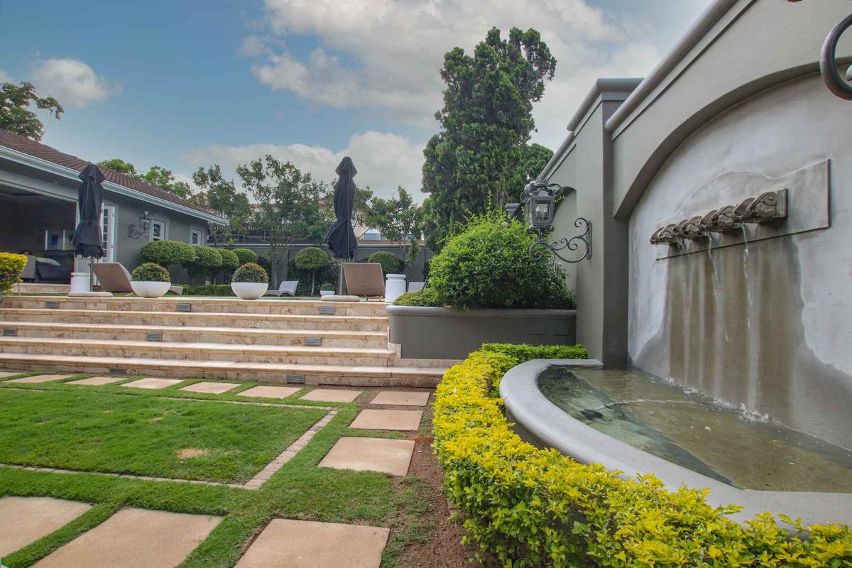 A beautifully landscaped rear garden featuring a tiered stone patio, manicured hedges, and a decorative wall-mounted water fountain.