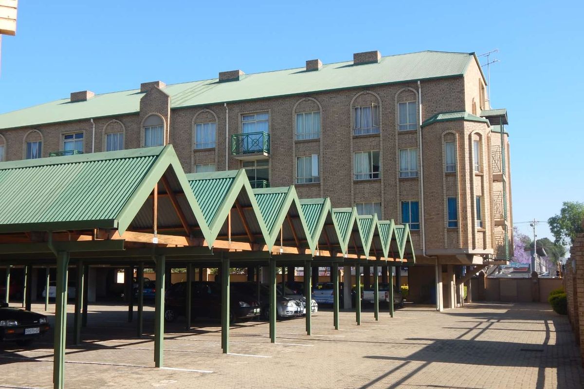 Exterior view of a multi-story brick apartment building featuring a green corrugated metal roof and a series of covered parking bays.