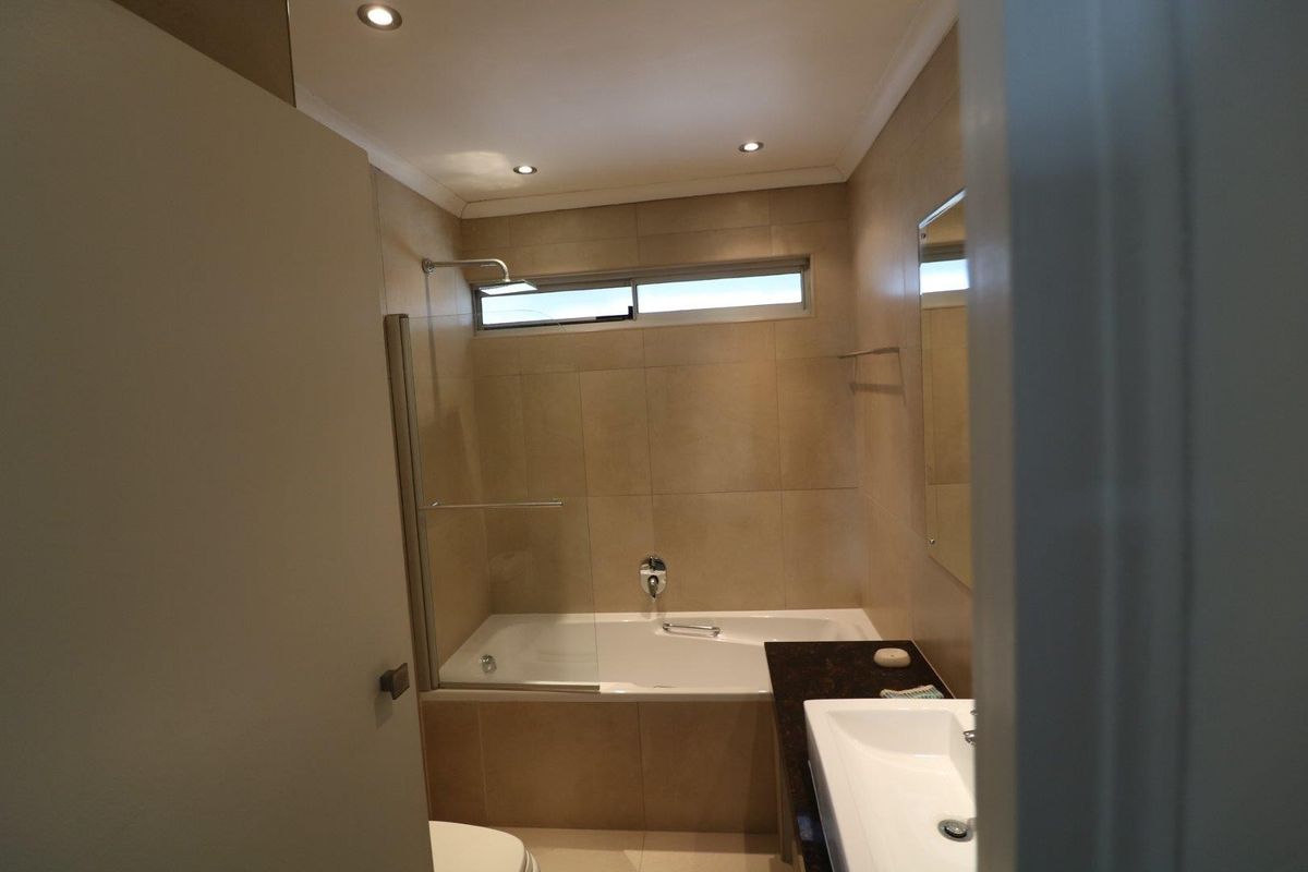 A modern bathroom featuring a built-in bathtub with a glass shower screen, a vanity basin with a granite countertop, and neutral wall tiling.