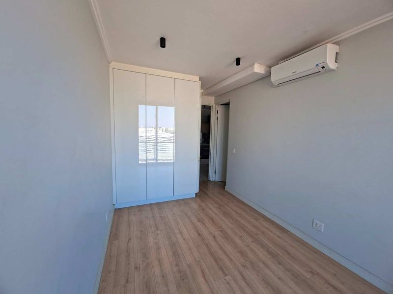 A bright, modern bedroom featuring light wood-look laminate flooring, a sleek white built-in wardrobe, and a wall-mounted split-system air conditioning unit.