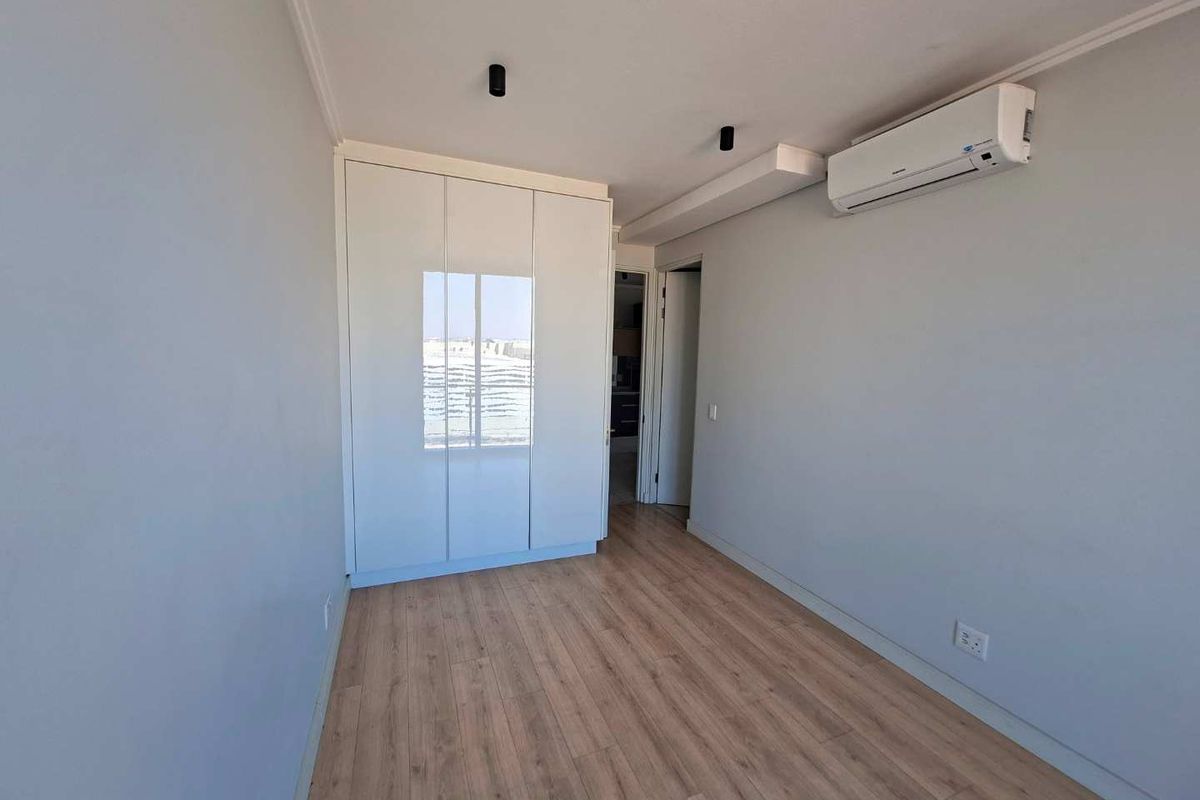 A bright, modern bedroom featuring light wood-look laminate flooring, a sleek white built-in wardrobe, and a wall-mounted split-system air conditioning unit.