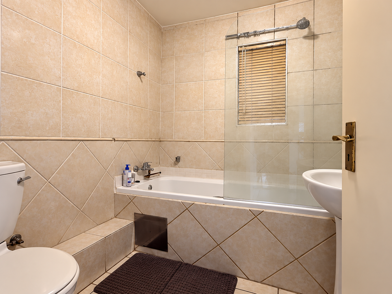 A functional bathroom featuring neutral ceramic wall and floor tiles, a built-in bathtub with a glass shower screen, and a pedestal sink.