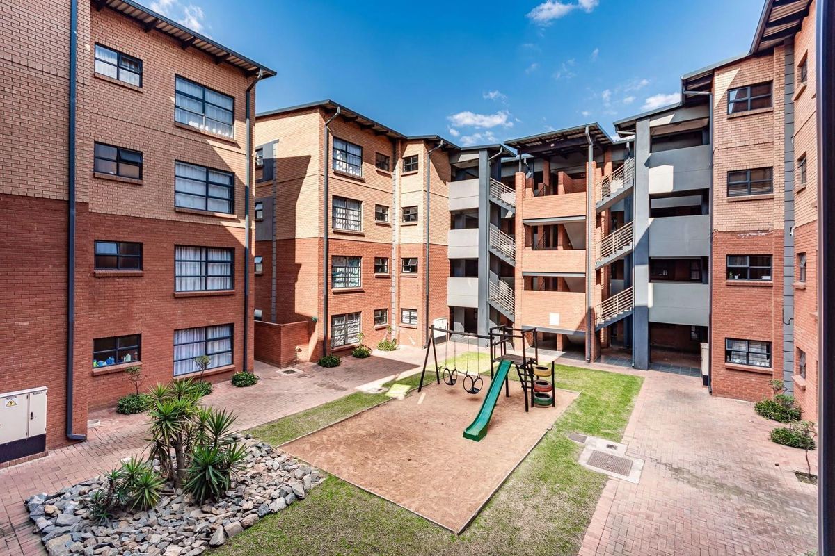 A view of the apartment complex courtyard featuring a paved area, landscaped garden, and a children's playground set between the brick building blocks.