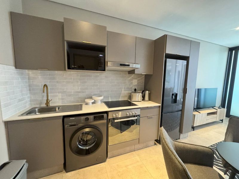 Modern open-plan kitchen featuring sleek grey cabinetry, stone countertops, subway tile backsplash, and integrated appliances including a washing machine and oven.