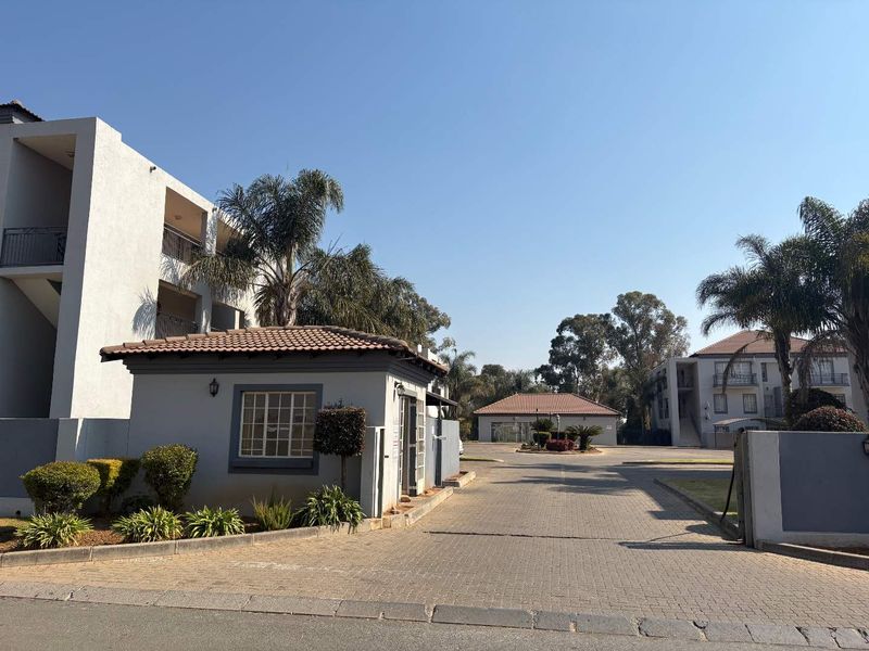 View of the apartment complex entrance featuring a security guard house, paved driveway, and landscaped grounds under a clear blue sky.