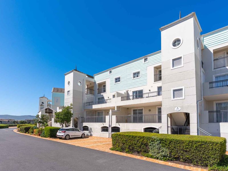 Exterior view of a multi-story apartment complex featuring stucco facades, private balconies with metal railings, and a paved driveway with landscaped hedges.