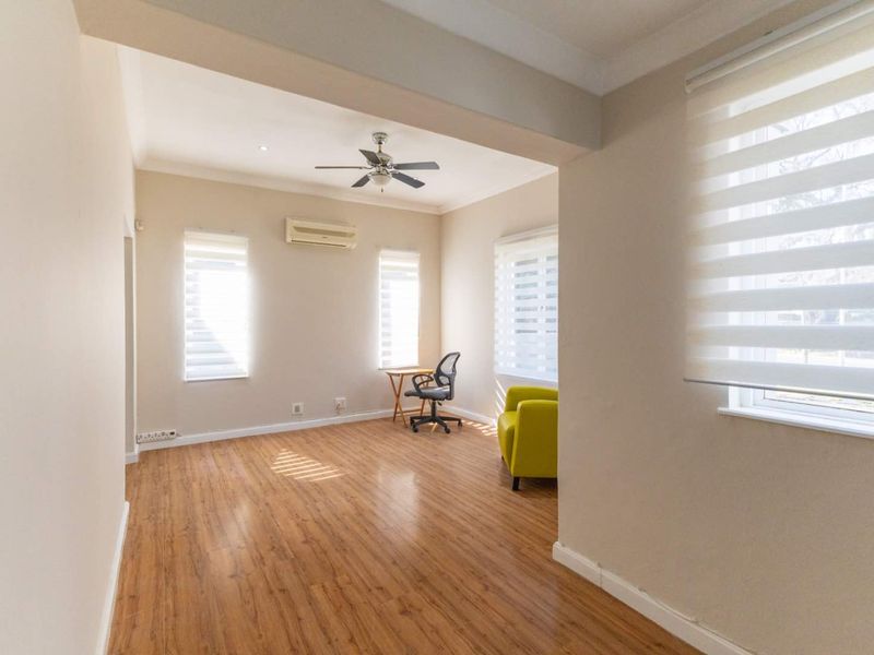 A bright living area featuring laminate flooring, a ceiling fan, and a wall-mounted air conditioning unit with modern roller blinds.