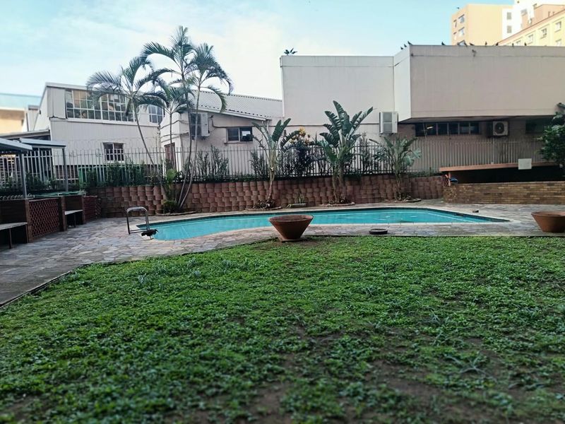 Communal outdoor pool area featuring a swimming pool, stone paving, and landscaped lawn, enclosed by a metal perimeter fence.