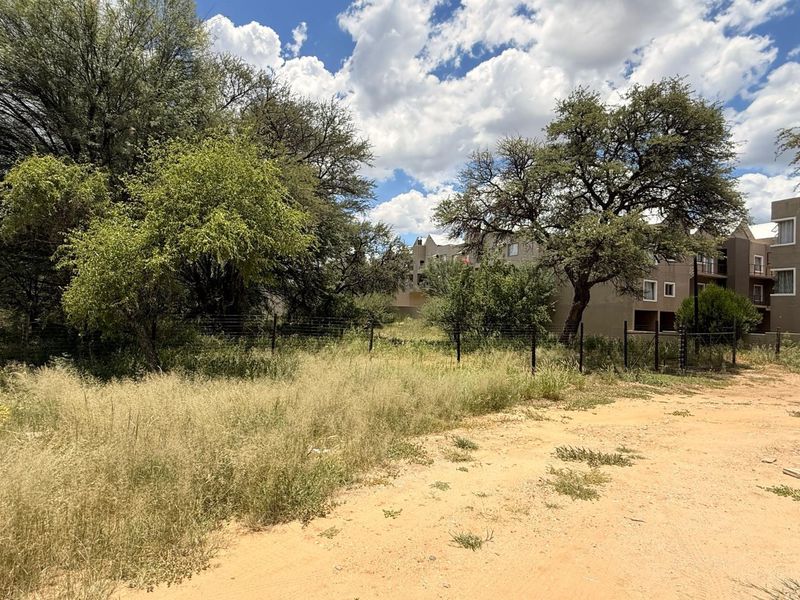 A vacant residential plot featuring natural vegetation, mature trees, and existing wire mesh fencing, adjacent to a multi-story residential complex.