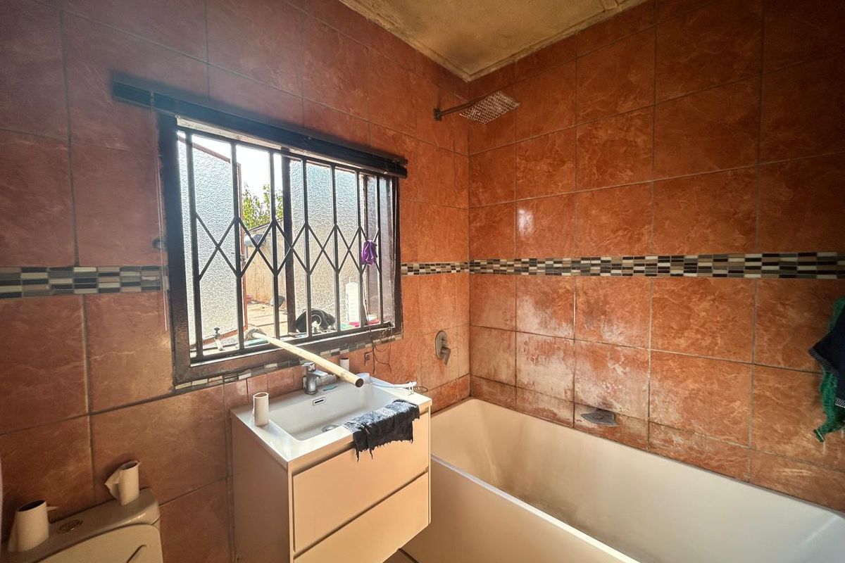 A dated bathroom featuring full-height orange wall tiles, a built-in bathtub, a wall-mounted vanity basin, and a window with security bars.
