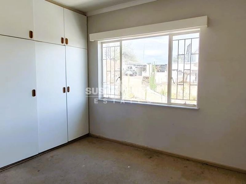 An empty bedroom featuring built-in cupboards and a window with security bars, requiring cosmetic updates to the flooring and walls.