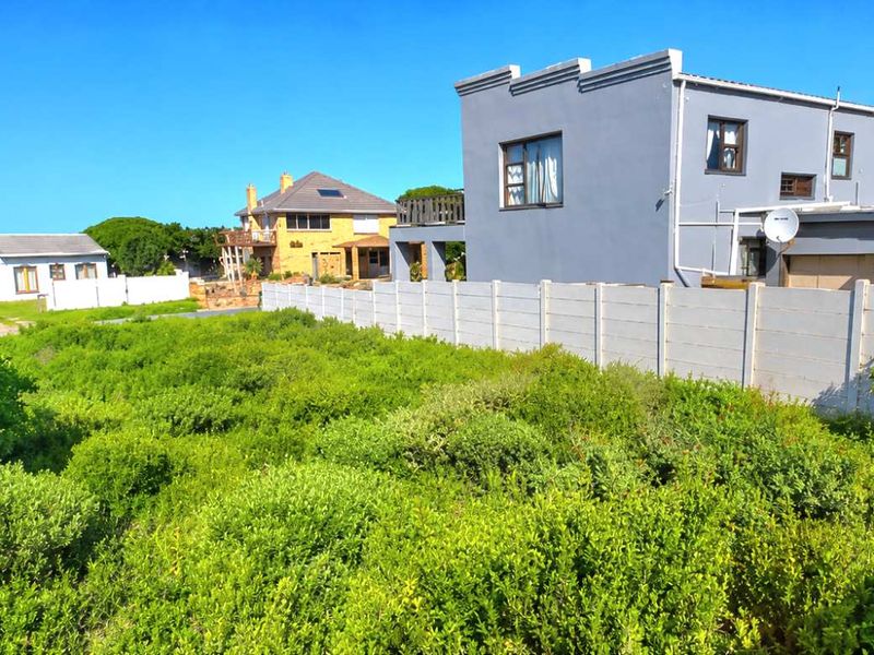 A vacant residential plot covered in natural vegetation, bordered by a precast concrete wall with neighboring residential properties visible in the background.