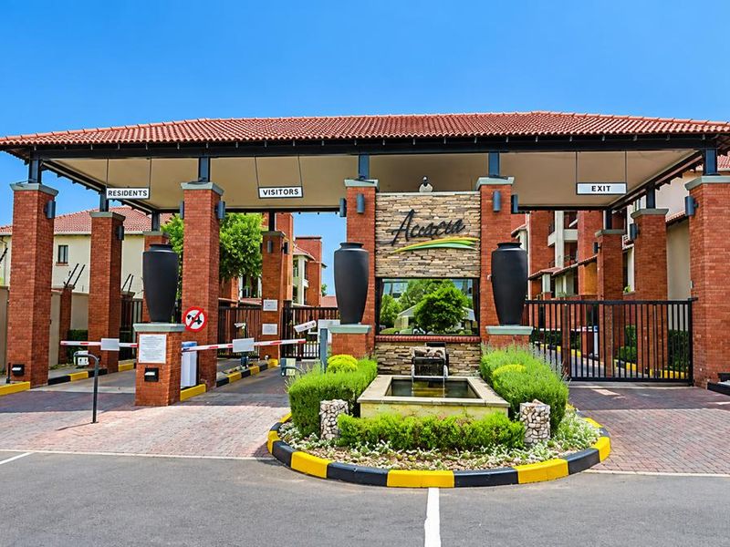 The secure entrance gatehouse of the Acacia complex, featuring brick pillars, a tiled roof, security boom gates, and a landscaped water feature.