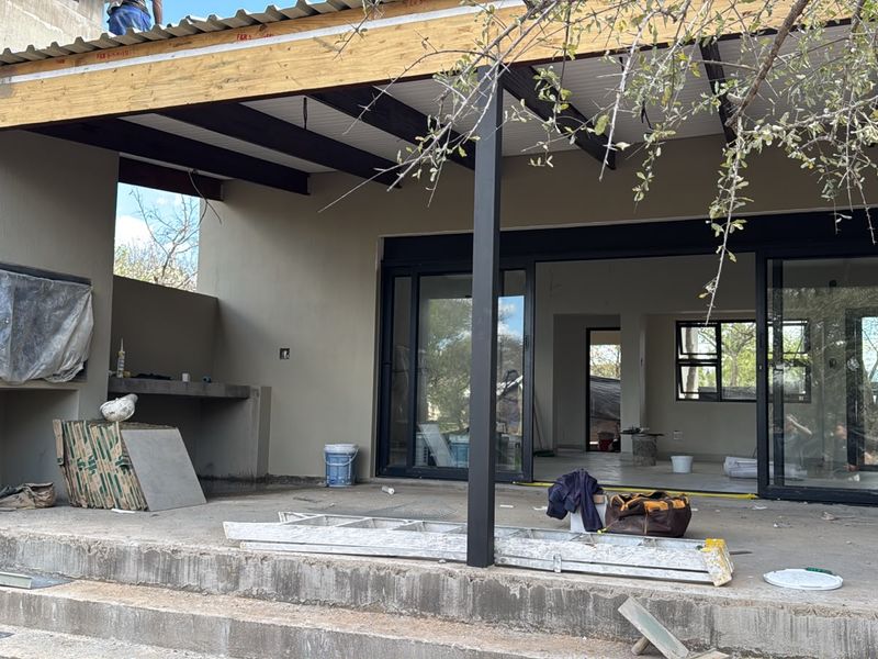 An unfinished outdoor patio area featuring a built-in braai, exposed ceiling beams, and large sliding glass doors leading into the main living space.