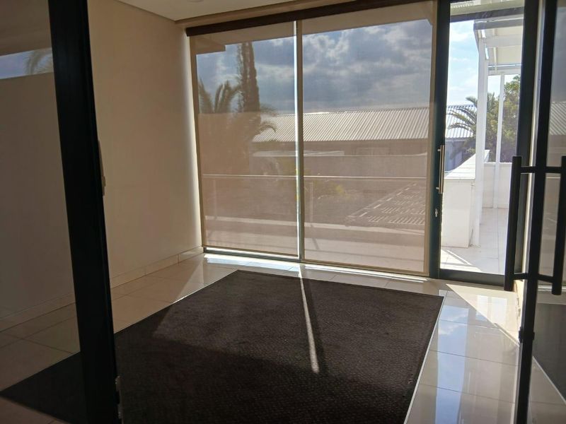 A bright, empty office space featuring large sliding glass doors with roller blinds, tiled flooring, and access to an outdoor balcony area.