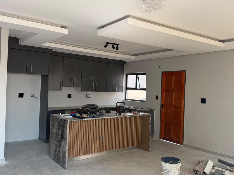 Modern kitchen featuring dark grey cabinetry, a stone-topped island with slatted wood detailing, and a contemporary dropped ceiling design, currently in final stages of completion.