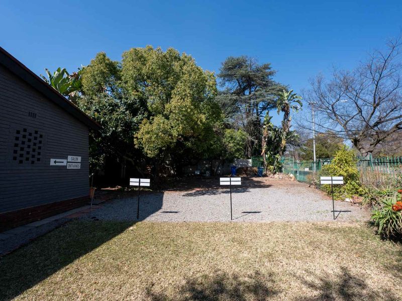 A rear outdoor area featuring a gravel parking space, a small lawn, and established trees, enclosed by metal perimeter fencing.