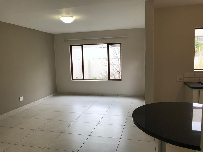A bright and spacious open-plan living area featuring neutral walls, large windows, and tiled flooring, seamlessly connected to a kitchen breakfast bar.