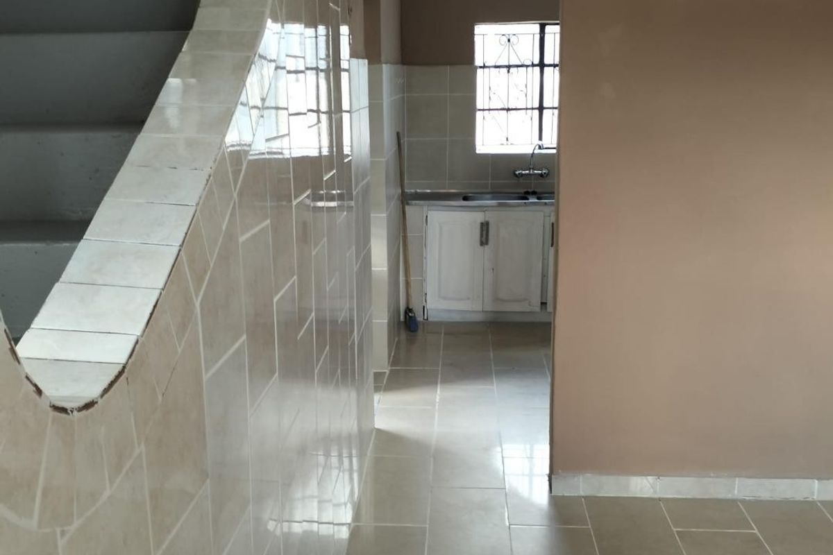 A view from a tiled hallway towards a kitchen area featuring built-in cabinetry, a sink, and a staircase with tiled wall finishes.