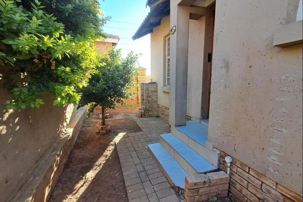 A side exterior view of the townhouse featuring a paved walkway, tiled entrance steps, and a brick boundary wall with established garden greenery.