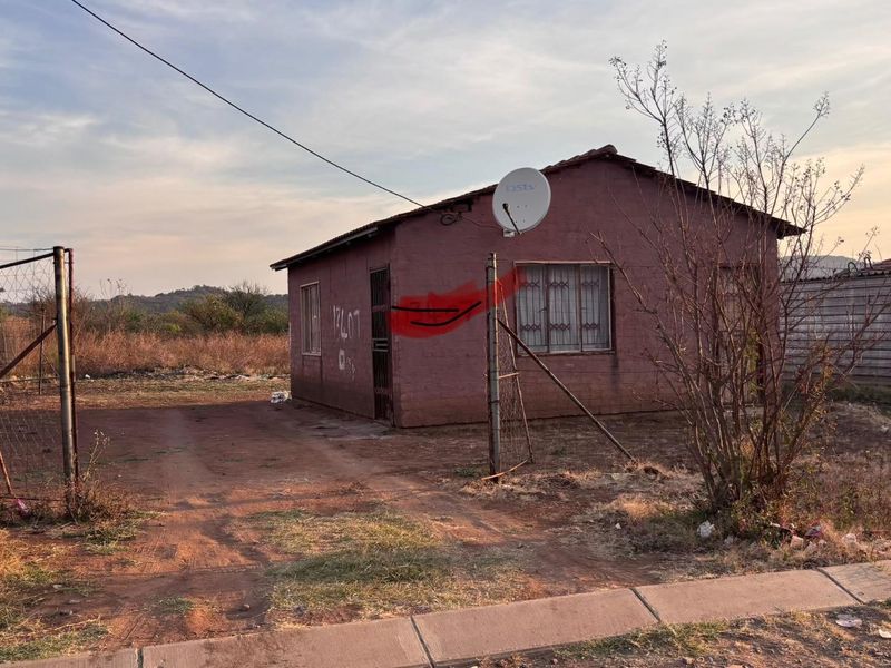 A detached single-story house featuring a tiled roof, security bars, and a satellite dish, situated on an unpaved plot with perimeter fencing.