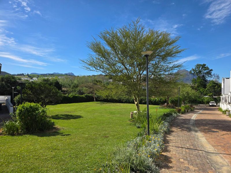 A well-maintained communal garden area featuring a paved walkway, mature trees, and landscaped lawn with mountain views.