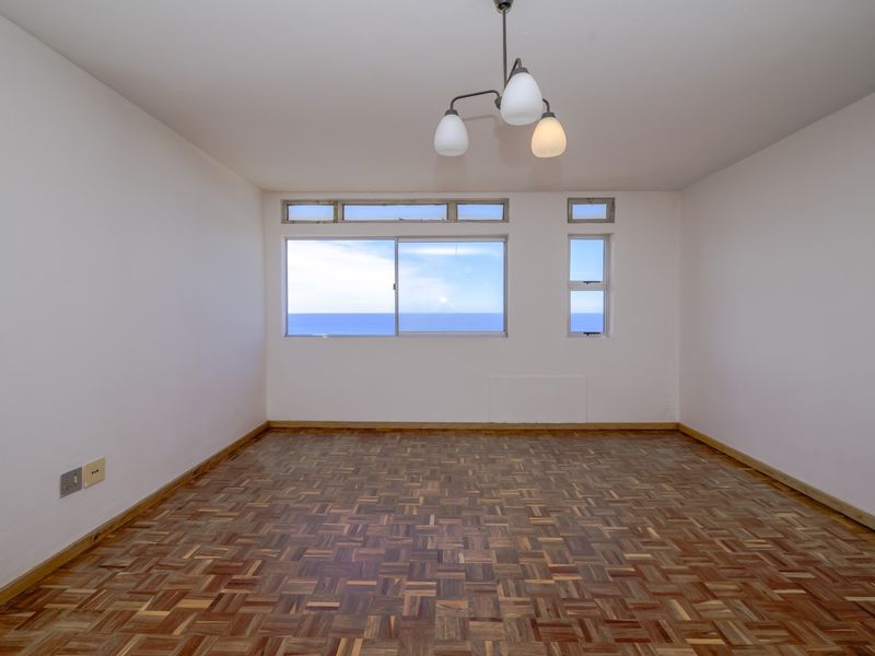 An empty living room featuring classic parquet flooring, large windows with ocean views, and a central ceiling light fixture.