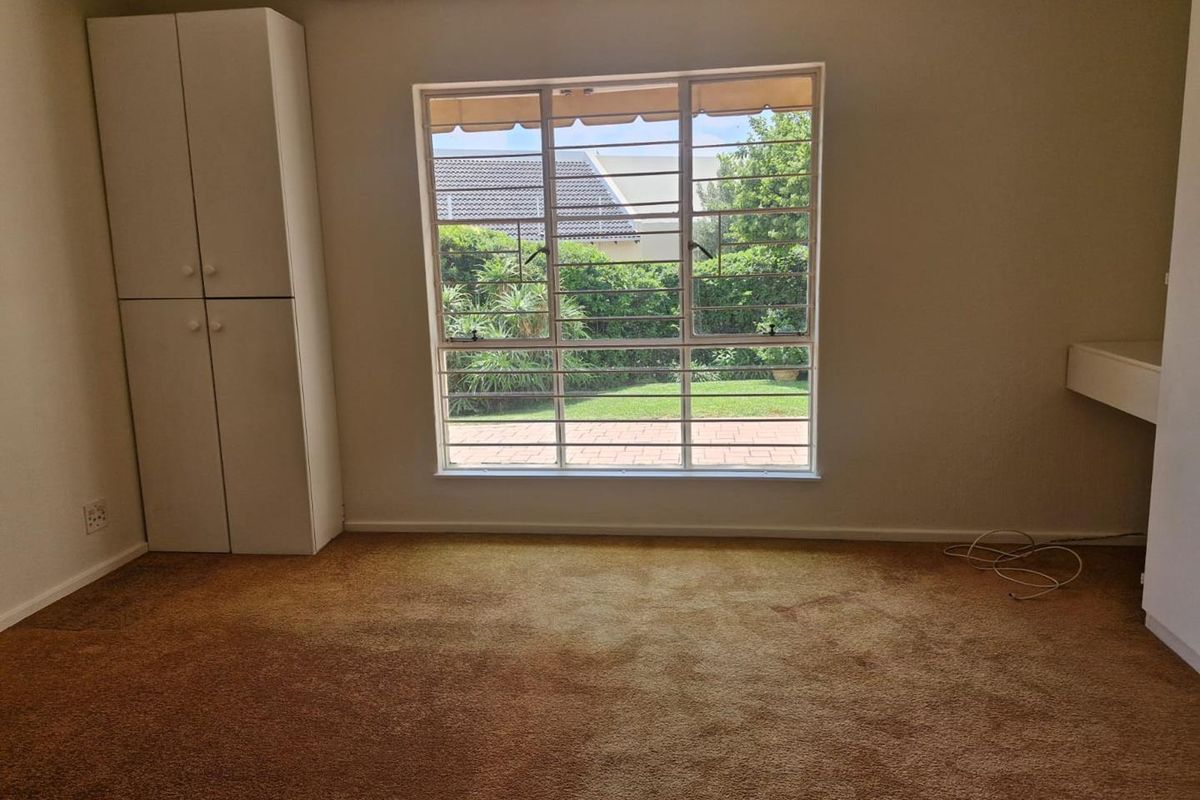 A bedroom featuring built-in cabinetry, a large window with security bars, and carpeted flooring, overlooking a private garden area.