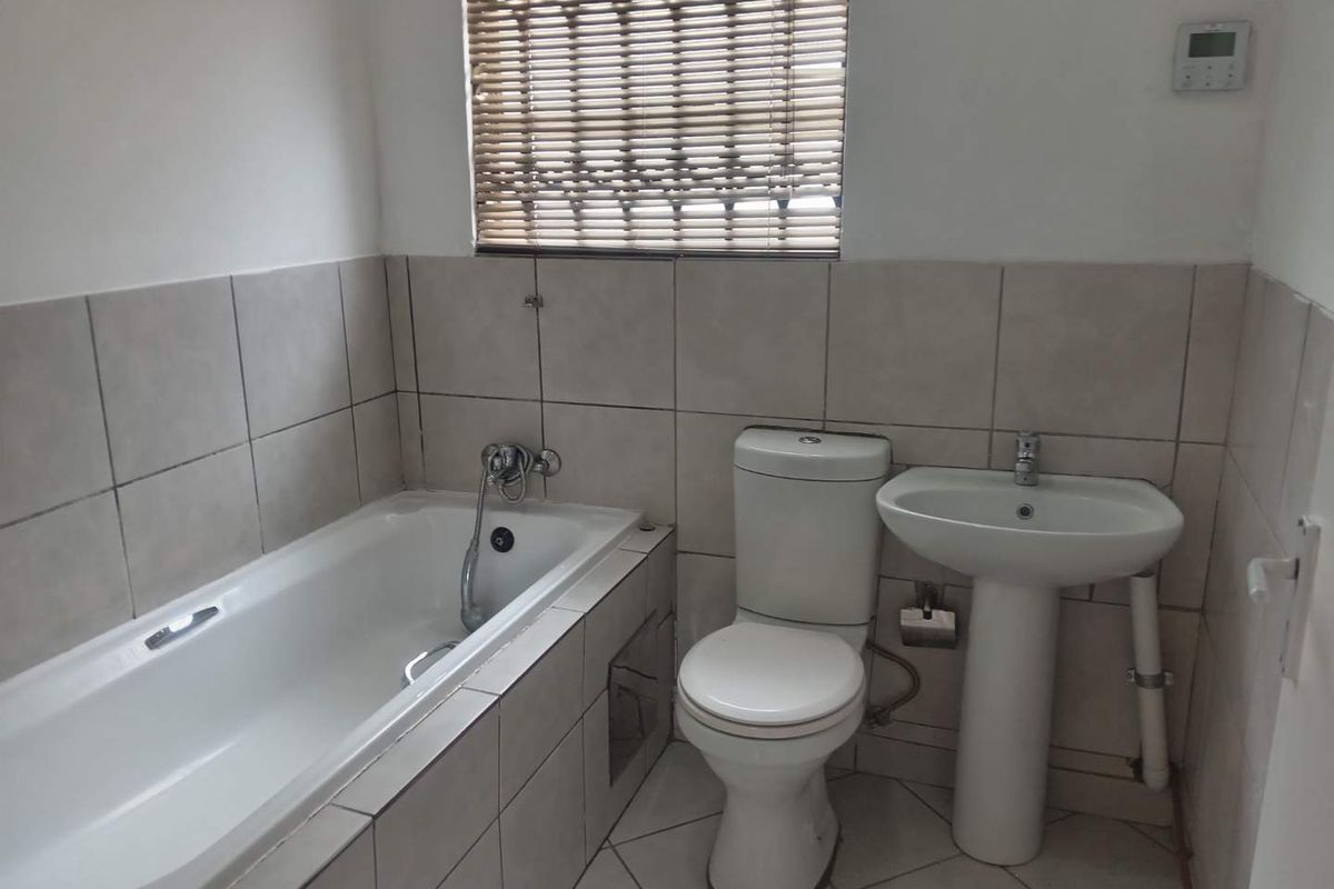 A functional bathroom featuring a standard bathtub, pedestal sink, and toilet with neutral wall tiling and a window with a bamboo blind.