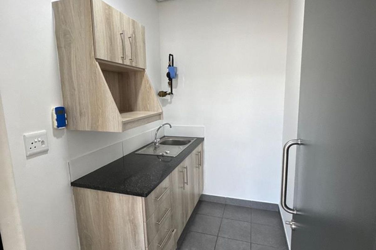 A compact office kitchenette featuring light wood-grain cabinetry, a dark stone countertop, a stainless steel sink, and modern grey floor tiling.