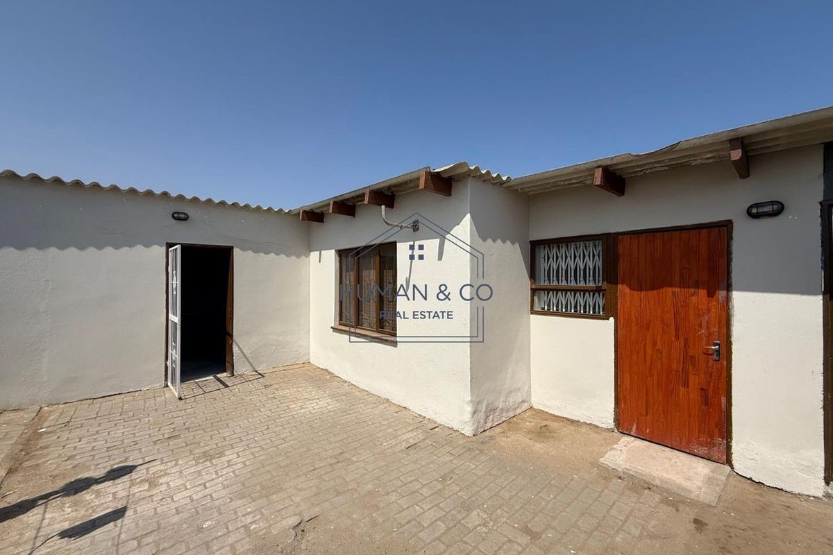 A rear exterior view of the property featuring a paved courtyard, wooden door, and security gates on windows.