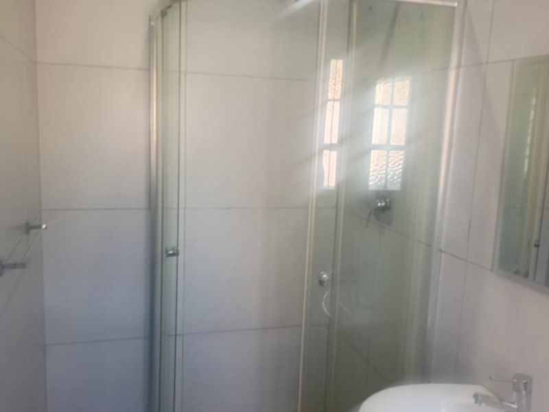 A functional bathroom featuring a curved glass shower enclosure, pedestal washbasin with chrome mixer tap, and neutral wall and floor tiling.
