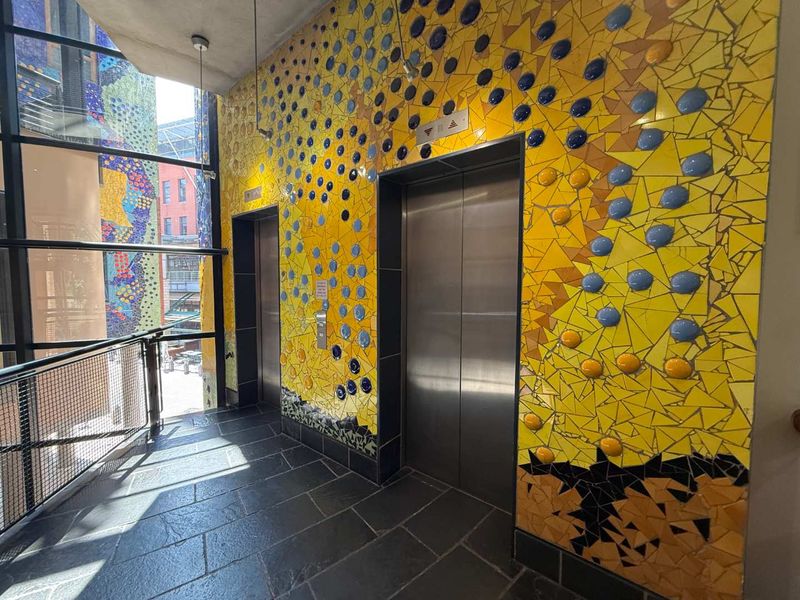 Modern office building elevator lobby featuring distinctive yellow and blue mosaic tile wall finishes, stainless steel elevator doors, and large glass windows.