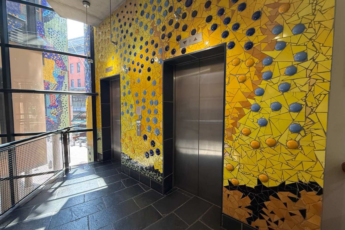 Modern office building elevator lobby featuring distinctive yellow and blue mosaic tile wall finishes, stainless steel elevator doors, and large glass windows.
