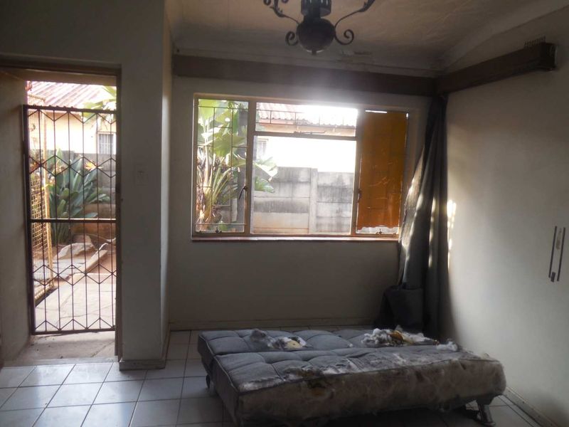 A sparsely furnished living area featuring tiled flooring, a large window, and direct access to an outdoor space through a security gate.